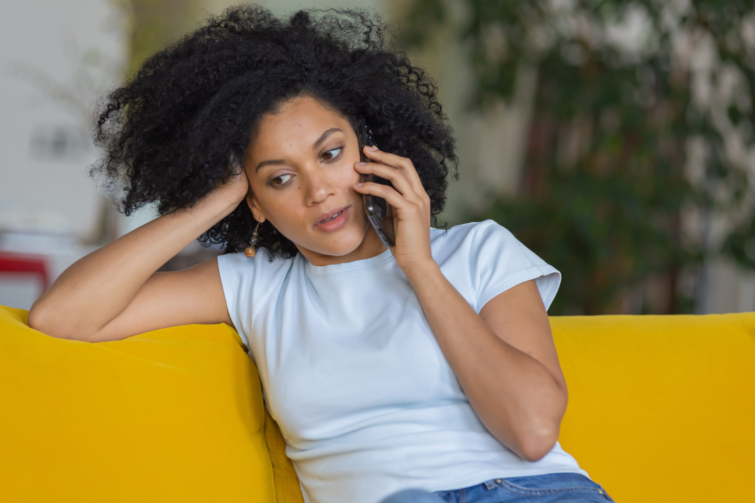 Portrait of a young African American woman talking for mobile phone. Brunette with curly hair sitting on yellow sofa in a bright home room. Close up.